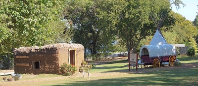 [In an open expanse with a few trees nearby stands a sod house on the left and a covered wagon in front of a tepee on the right.]